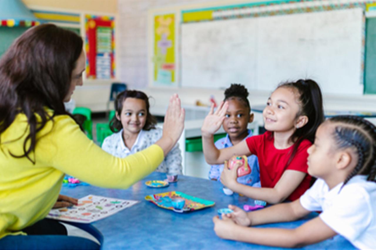 four young children students with a teacher wearing a yellow shirt high fiving inside an elementary school classroom 