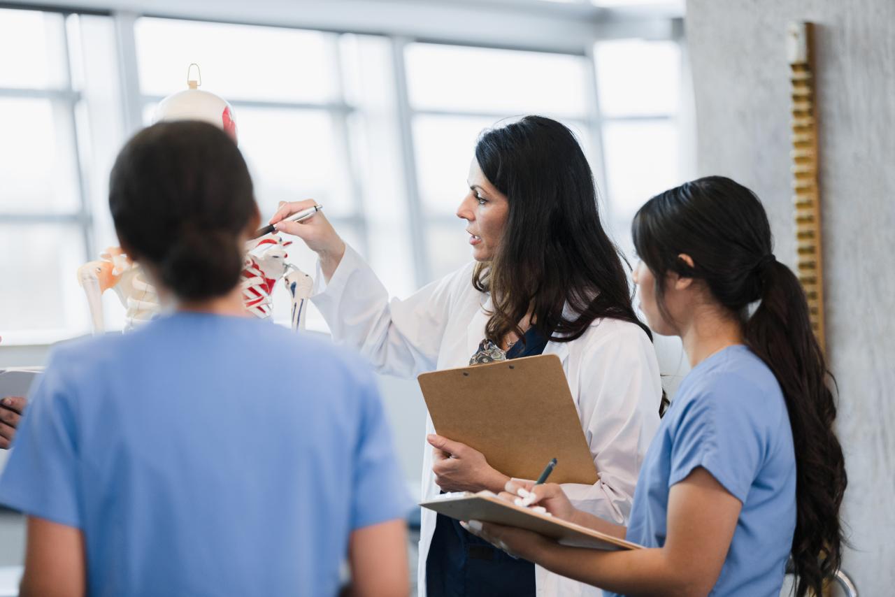 A teacher in a white lab coat points to a model skeleton while two students watch.