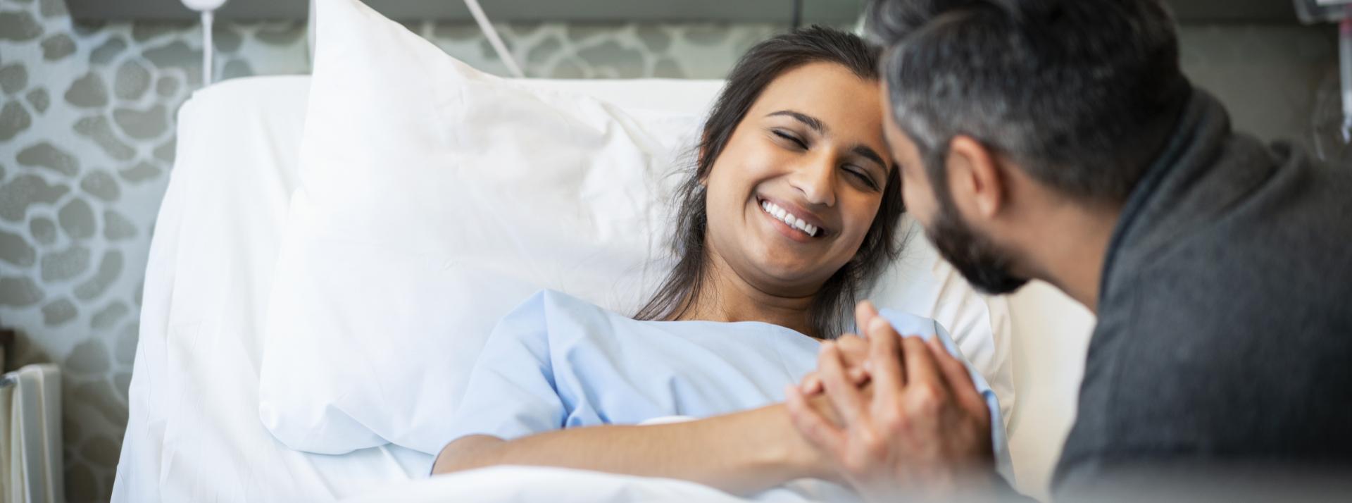 A person visiting someone in the hospital. The patient is lying in bed and smiling