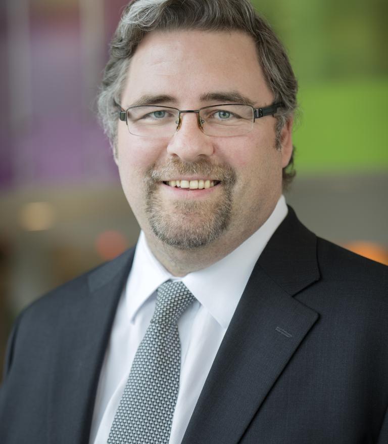 A portrait of William Duvall in front of a colourful background.  He is wearing a grey tie and dark blue suit.