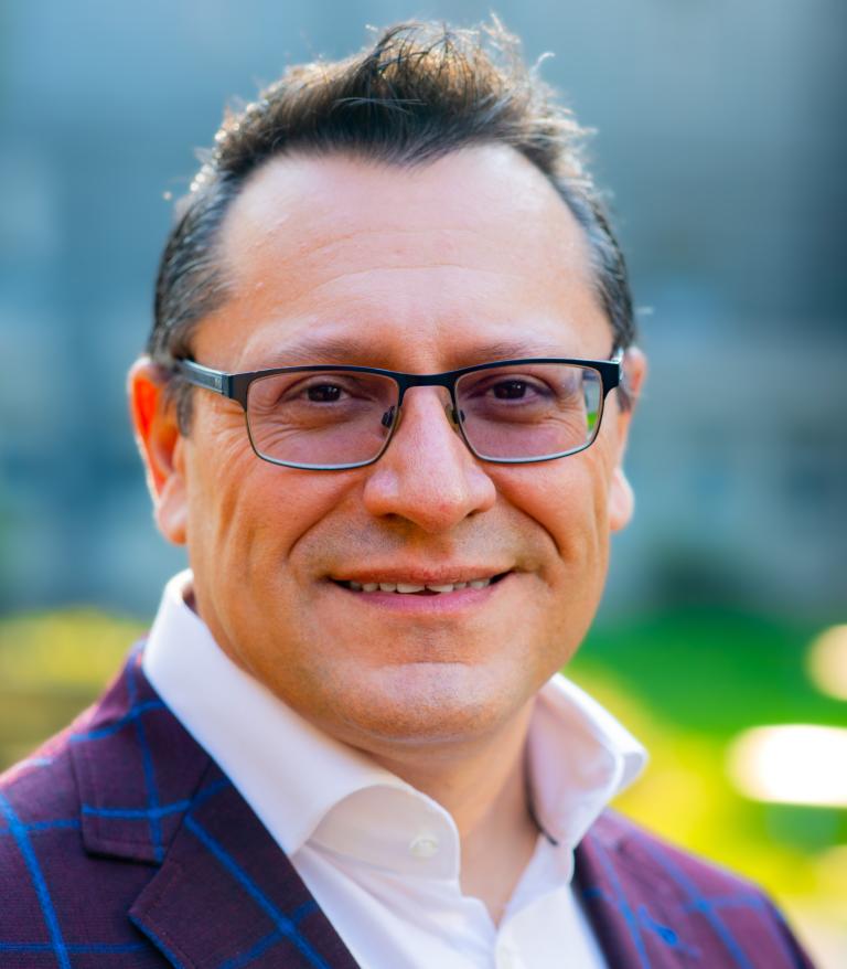 A portrait of Fernando Pica outside on a sunny day.  He is wearing a white shirt and purple blazer