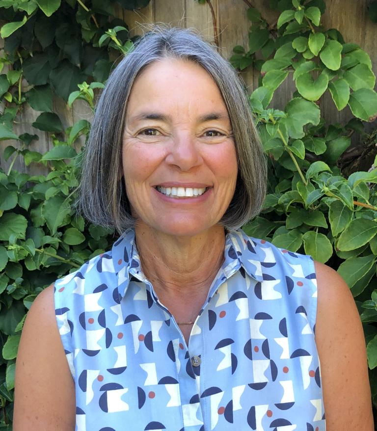 A portrait of Kathy Greenberg in front of green plants.  She is wearing a blue and white patterned top