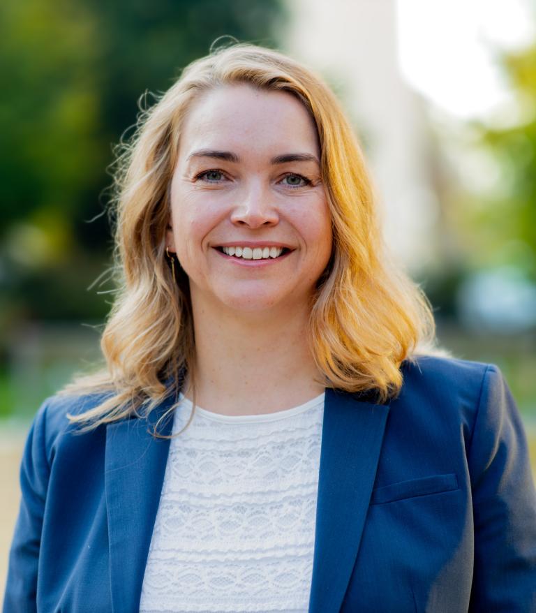 A portrait of Lorraine Blackburn outside on a sunny day.  She is wearing a white shirt and blue blazer.