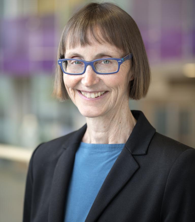 Portrait of Margaret McGregor in front of a colourful background, she is wearing a blue shirt and black blazer