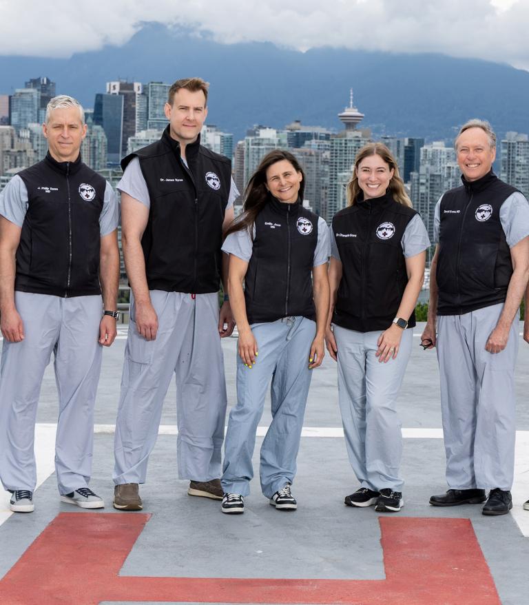 7 physicians wearing blue medical scrubs and black vests standing on a hospital rooftop with downtown Vancouver skyline and mountains in the background