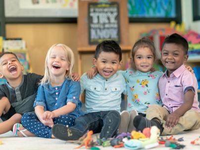 Children sitting in a line with their arms around each other. The kids are laughing and smiling directly at the camera.