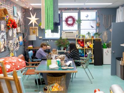Art Works Studio at G.F. Strong, with two people sitting at a table doing art. There are different kinds of art on the walls and hanging from the ceiling.