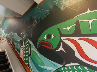An indigenous artwork mural on a wall going up alongside a long staircase. The artwork features large green salmon with black, white and red details over top of a river and black trees forest.