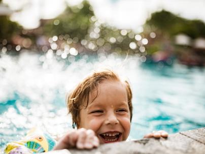 A happy child wearing water wings holding the edge of an outdoor pool on a sunny day.