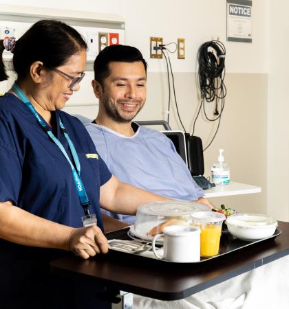 A food service staff member bringing a seated patient their meal in a hospital room.