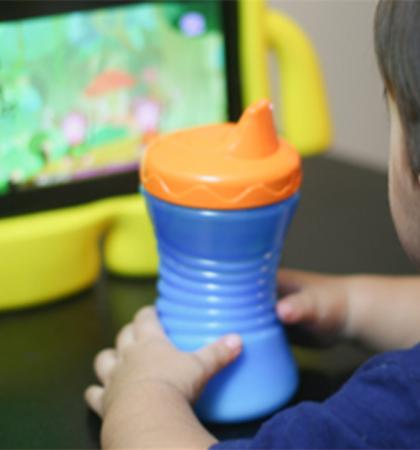 A toddler in blue shirt with a blue and orange drinking cup in front of a tablet with a yellow case