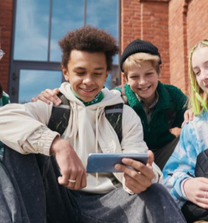 a group of four teens sitting outside a brick building looking at a smartphone screen